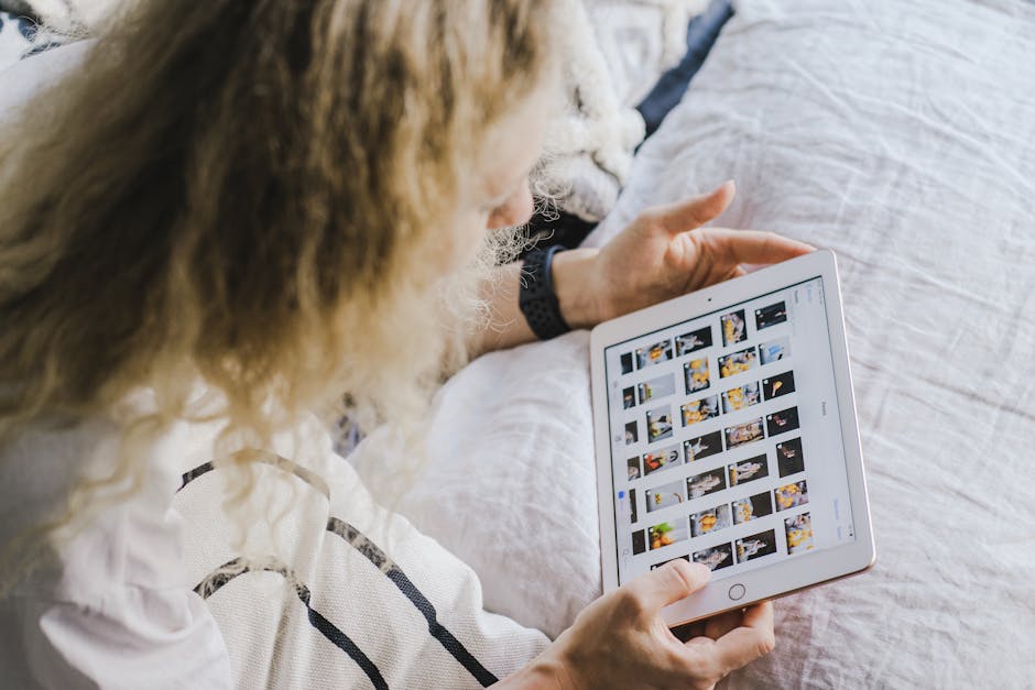 A woman sitting on a bed browsing images on a tablet with white bedding.