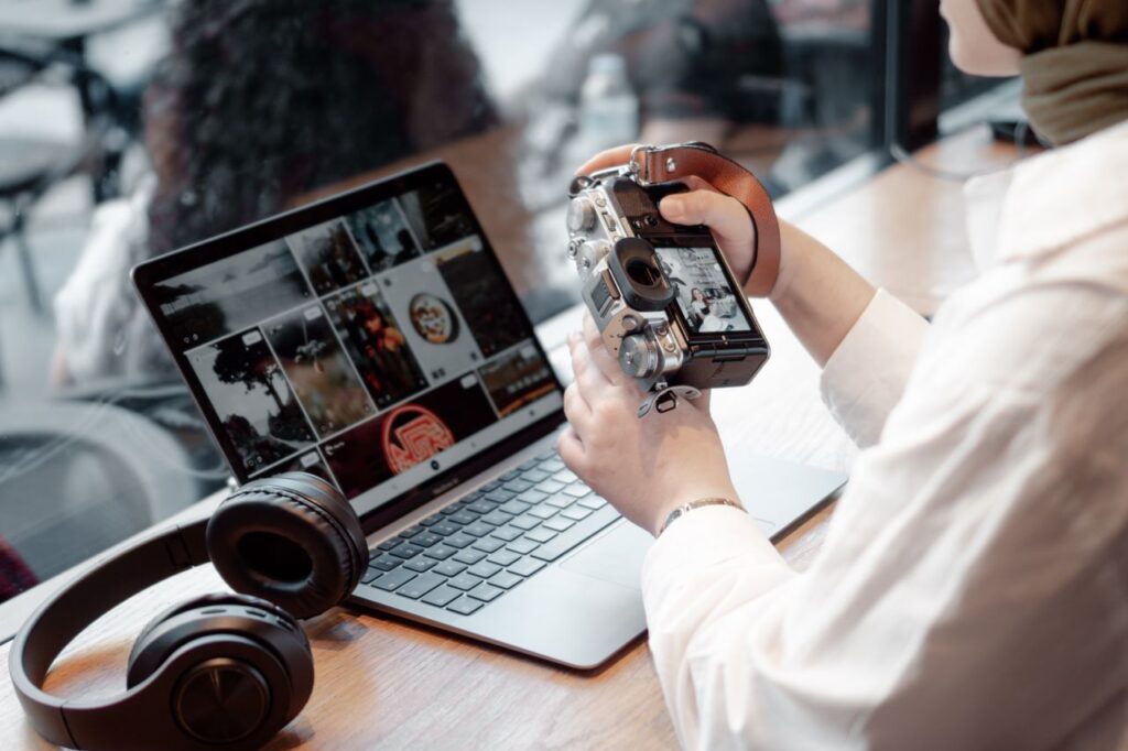 A woman editing photos on her laptop while holding a camera, with headphones nearby on a desk.